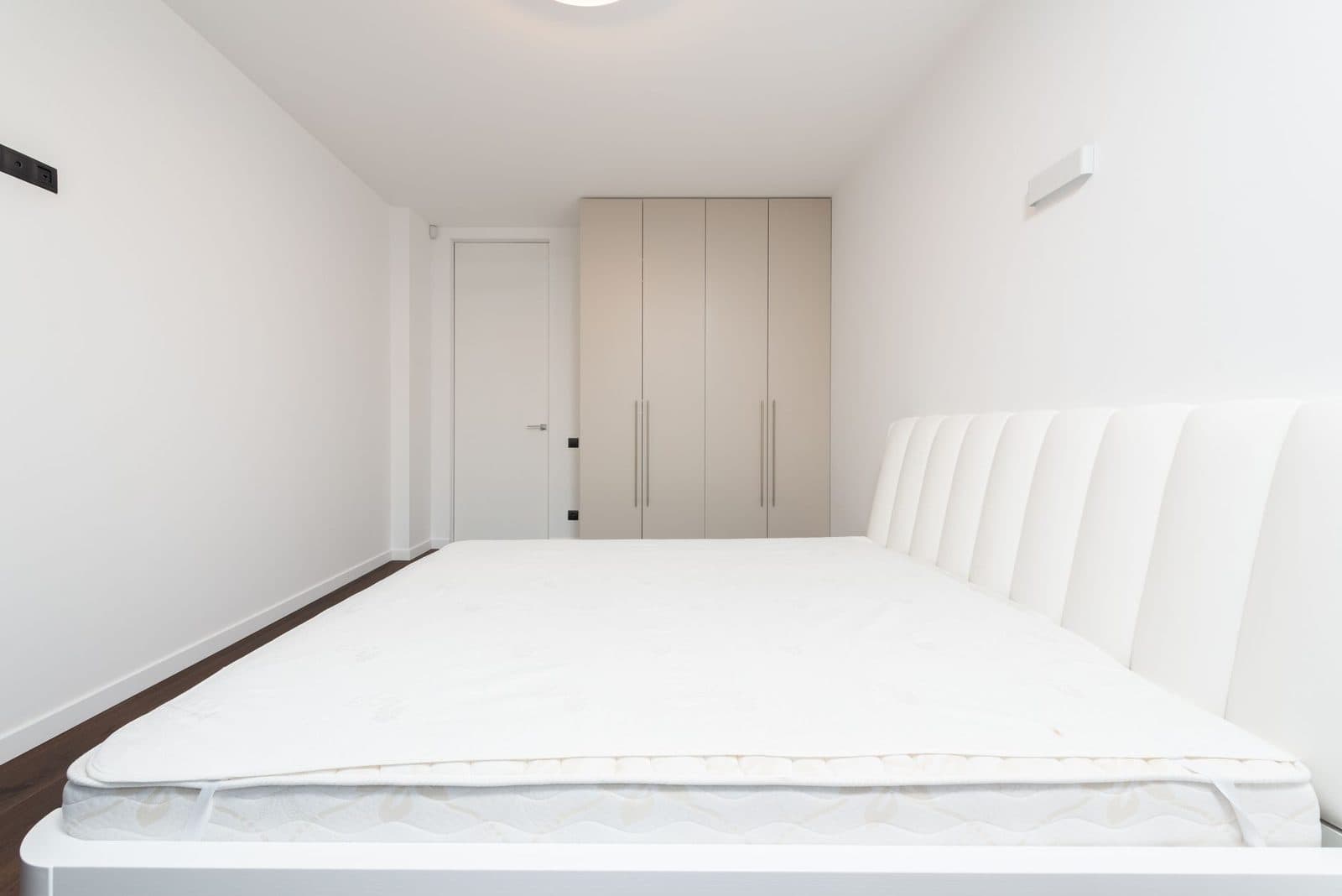 Colchón sin ropa de cama en una cama con cabecero blanco en un dormitorio luminoso con armario beis