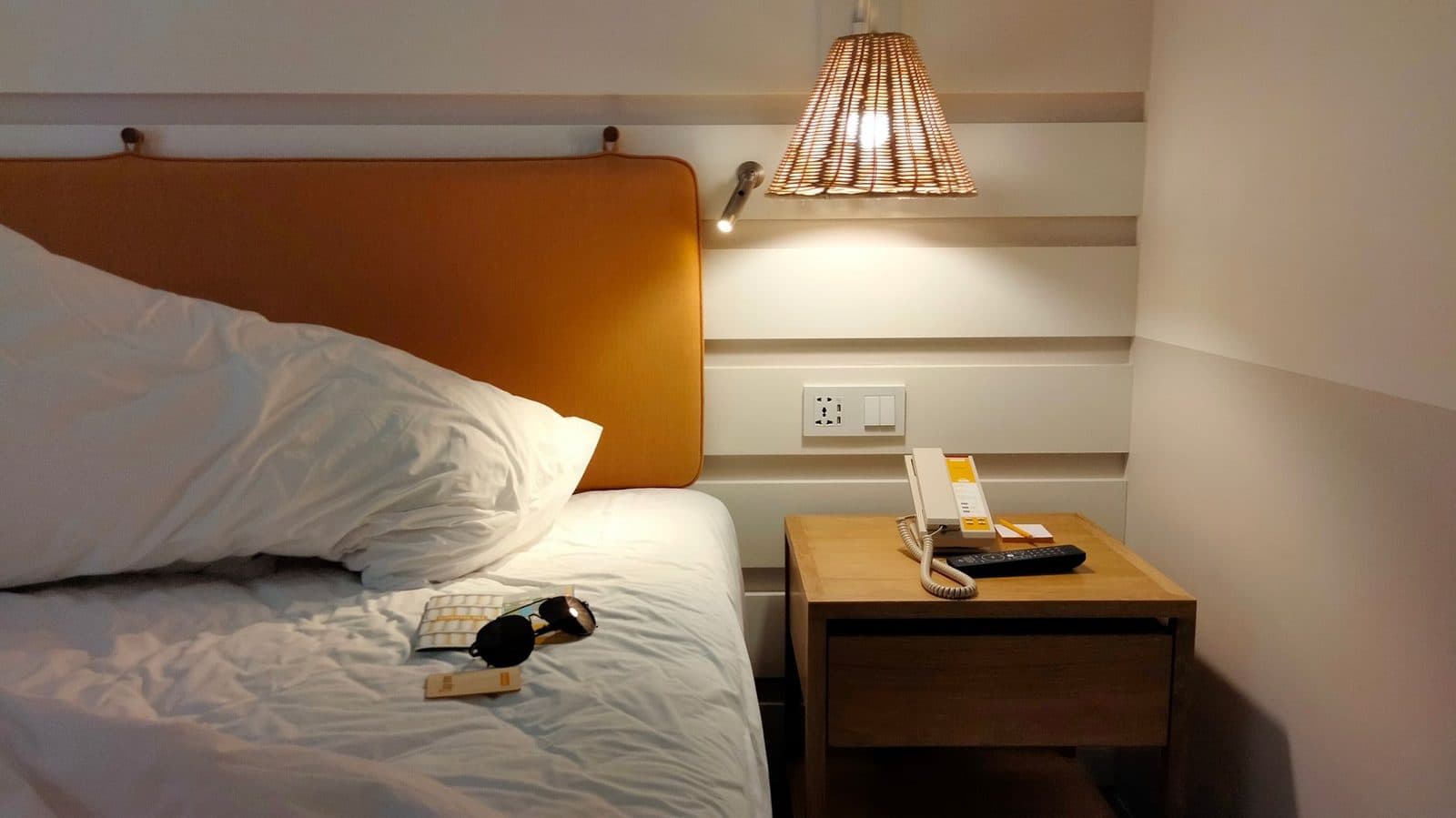 Lit défait aux draps blancs avec tête de lit en cuir, table de chevet en bois et lampe allumée