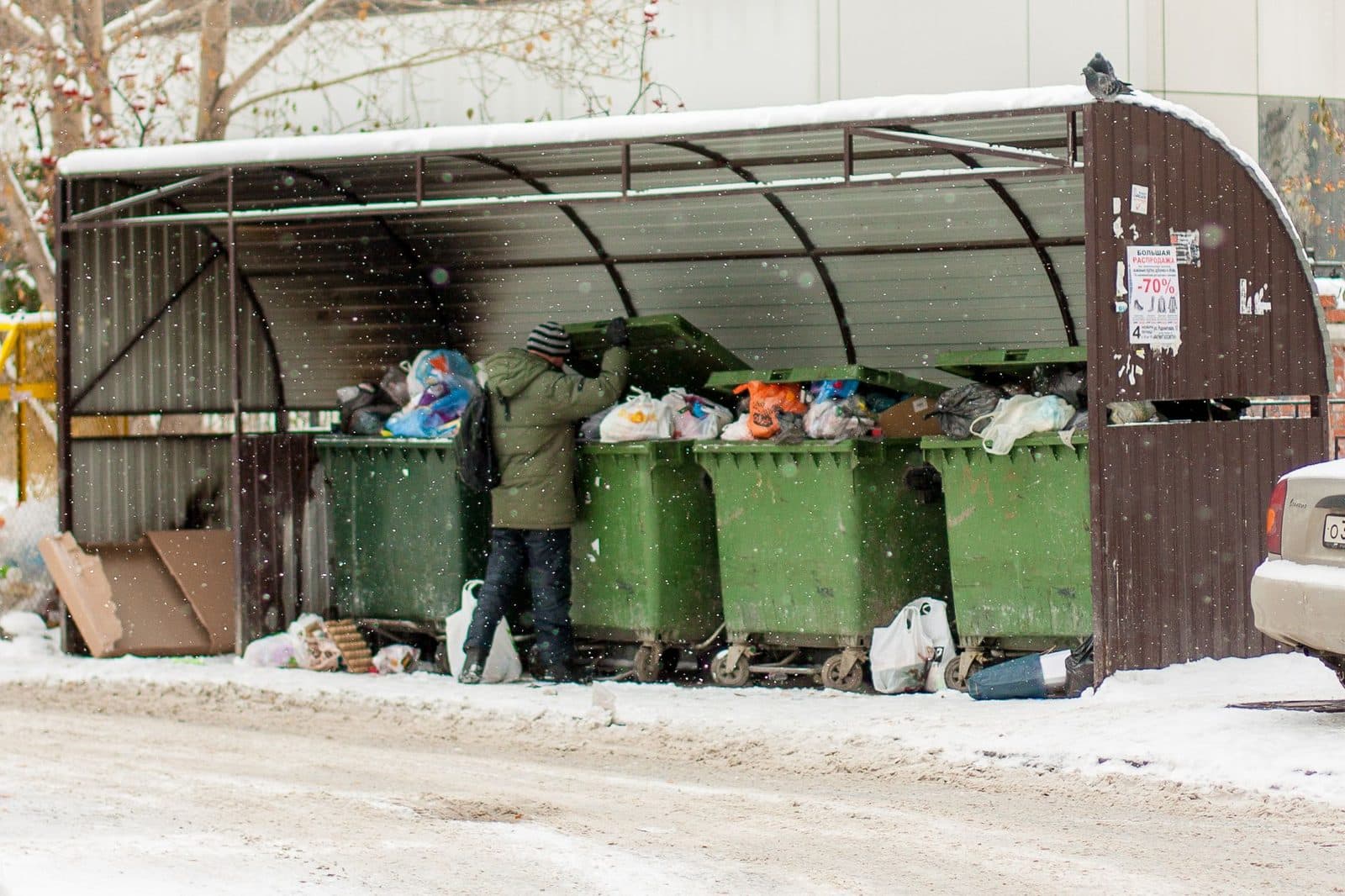 Abri à conteneurs à ordures débordant de sacs poubelle sous la neige, un homme fouille les bacs verts