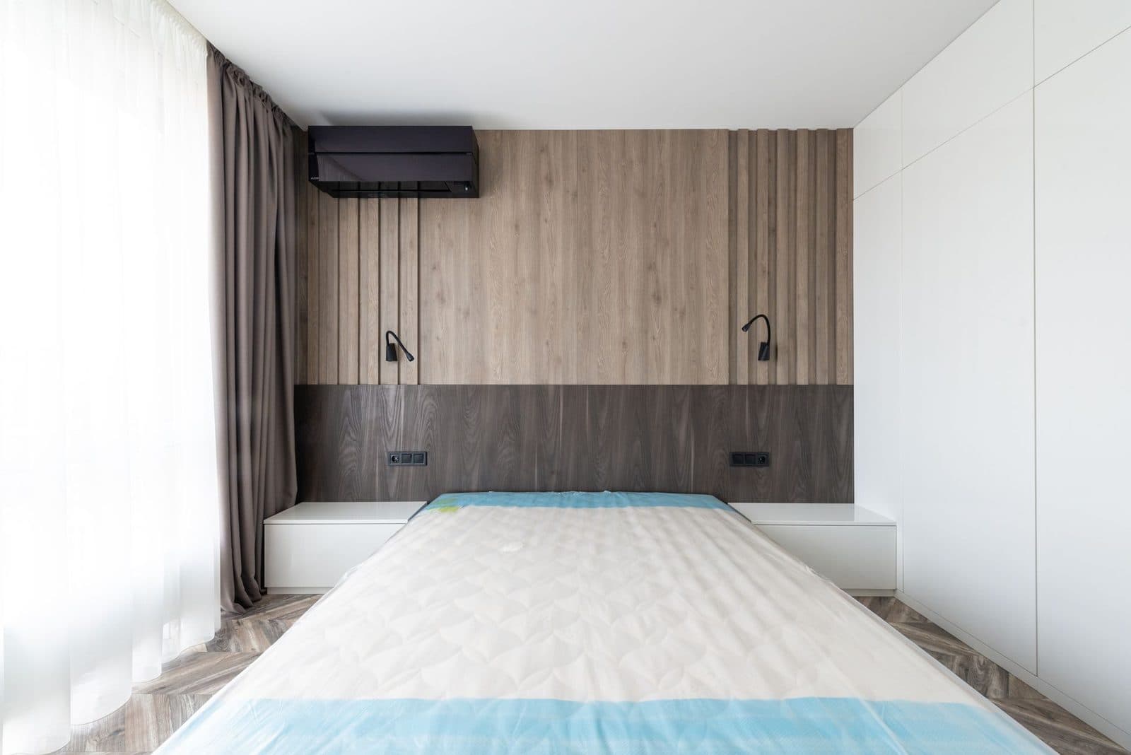 Chambre moderne avec matelas nu sur le lit, tête de lit en bois, liseuses et climatiseur mural