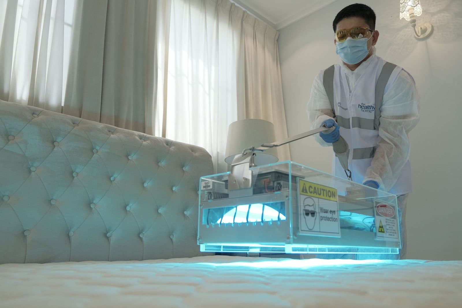 Techniker mit Maske behandelt eine Schlafzimmermatratze mit einem blauen UV-Lichtgerät mit Warnhinweisen