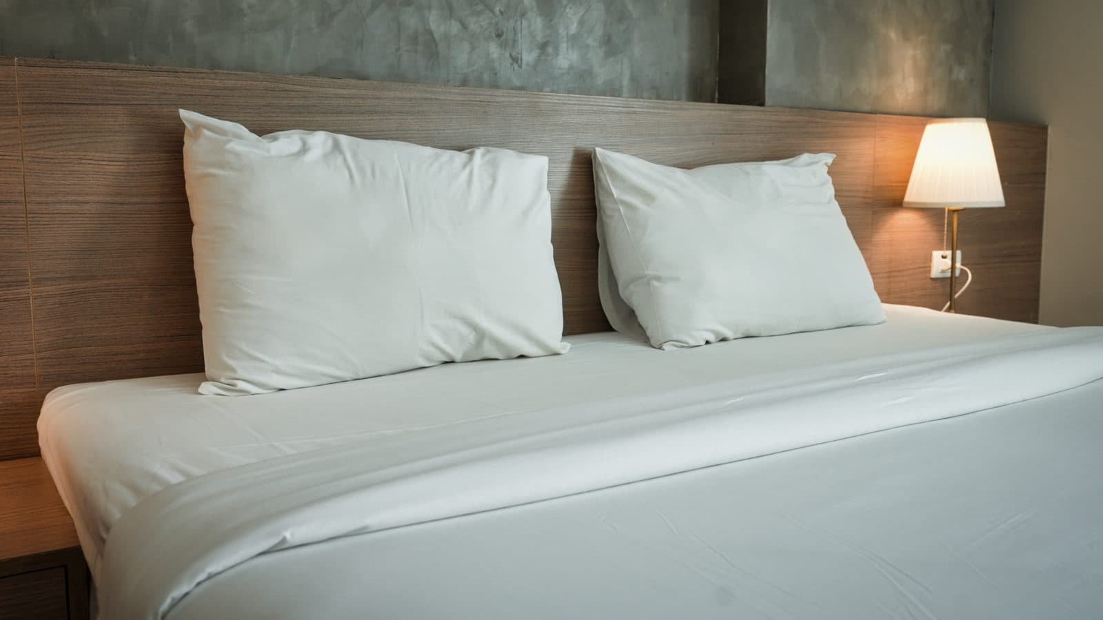 Lit double avec draps blancs et deux oreillers, tête de lit en bois et lampe de chevet allumée