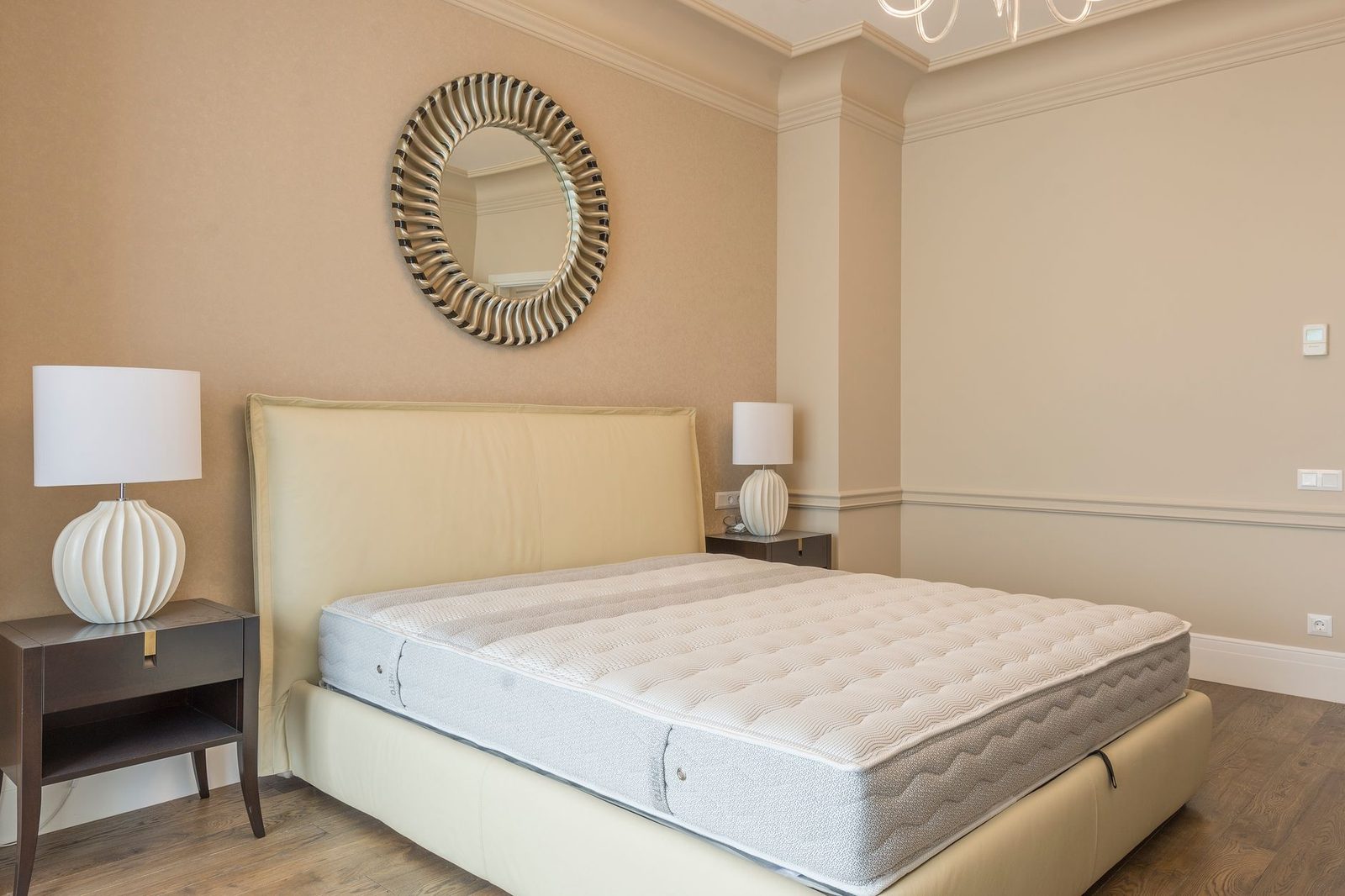 Slaapkamer met bed met kaal matras zonder lakens, beige hoofdeinde, nachtkastjes en ronde spiegel