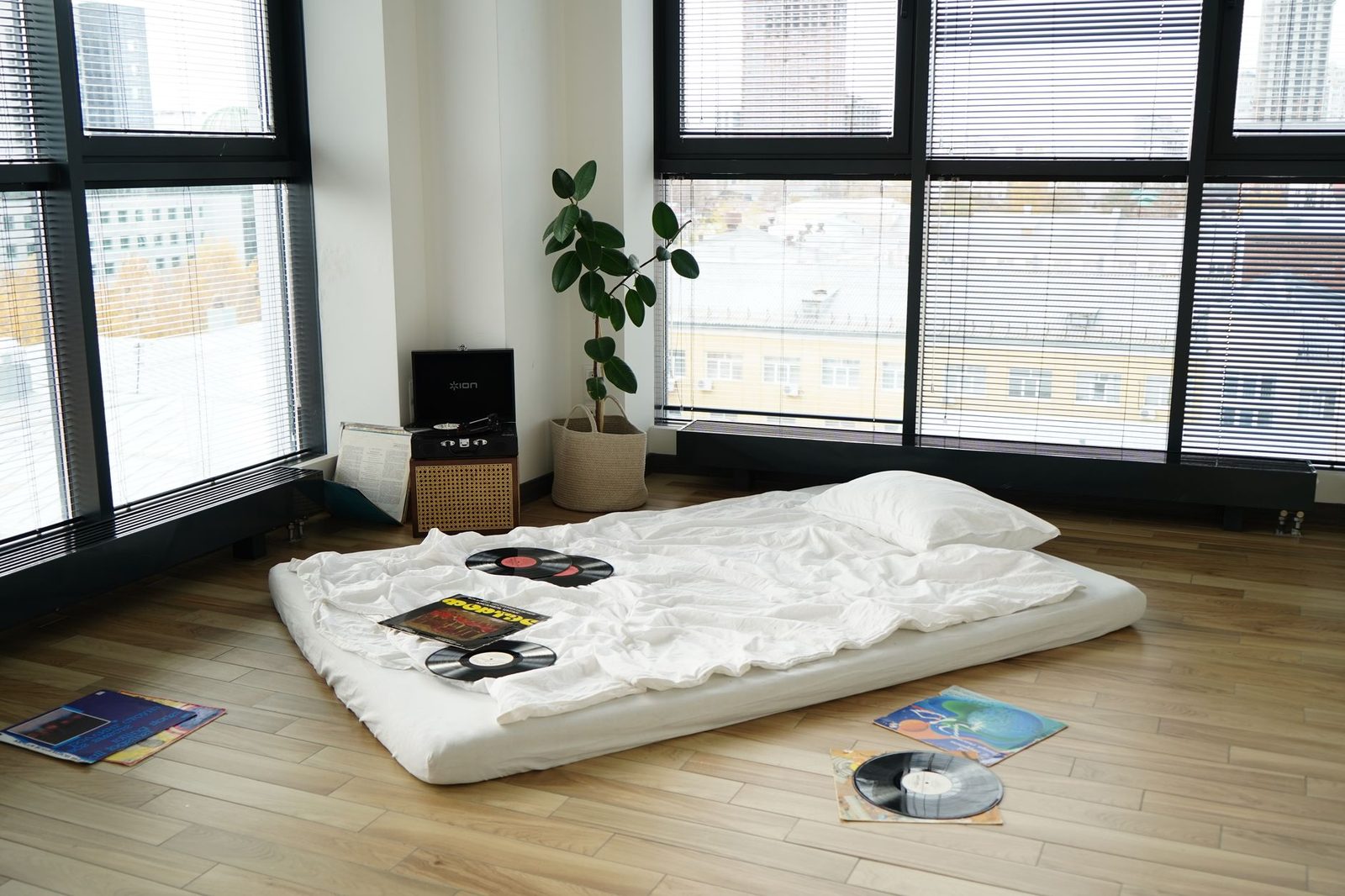 Matelas posé au sol avec draps blancs et oreiller dans une chambre lumineuse, disques vinyles autour
