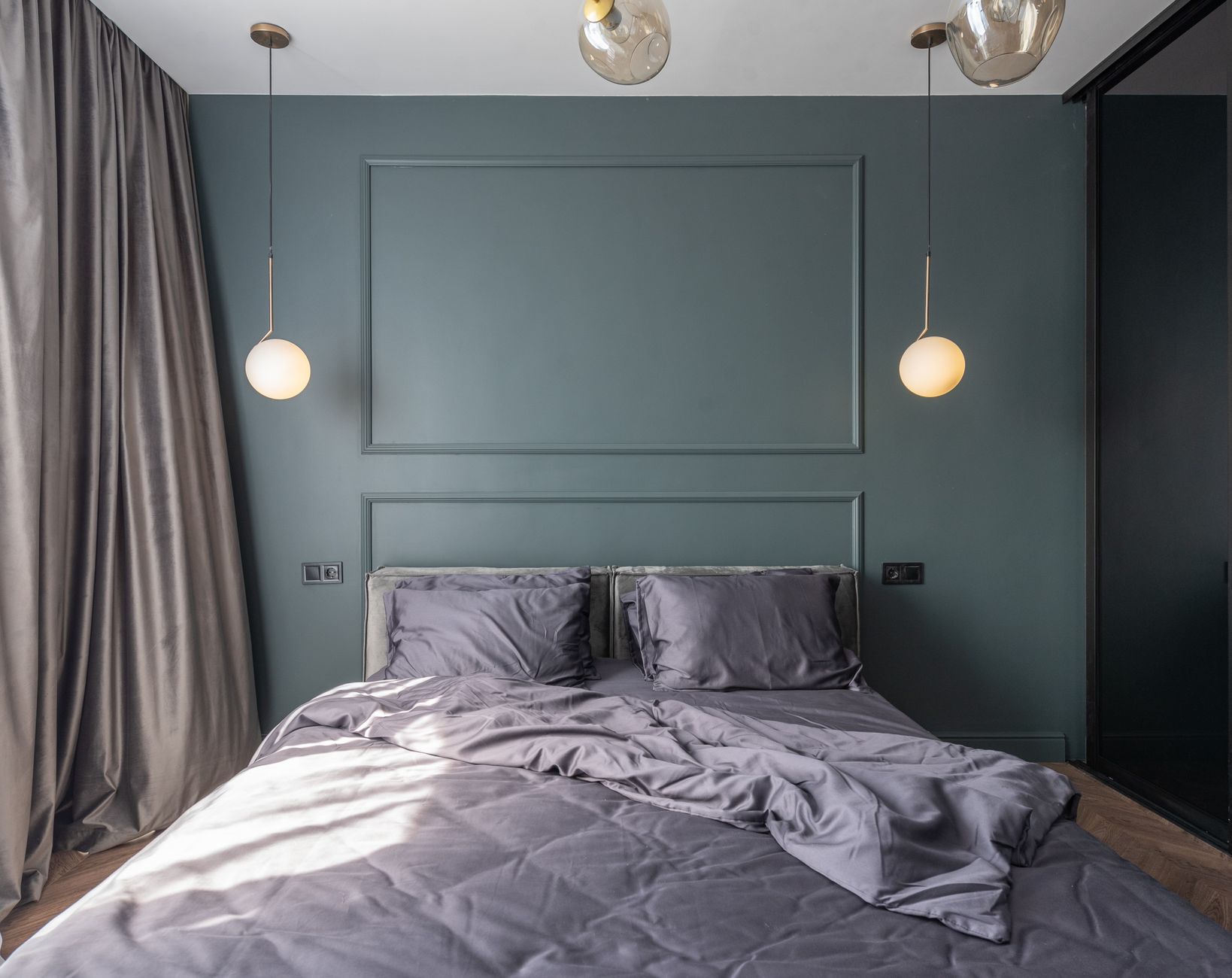 Zerwühltes Bett mit grauer Bettwäsche in einem Schlafzimmer mit graugrünen Wänden und leuchtenden Pendelleuchten