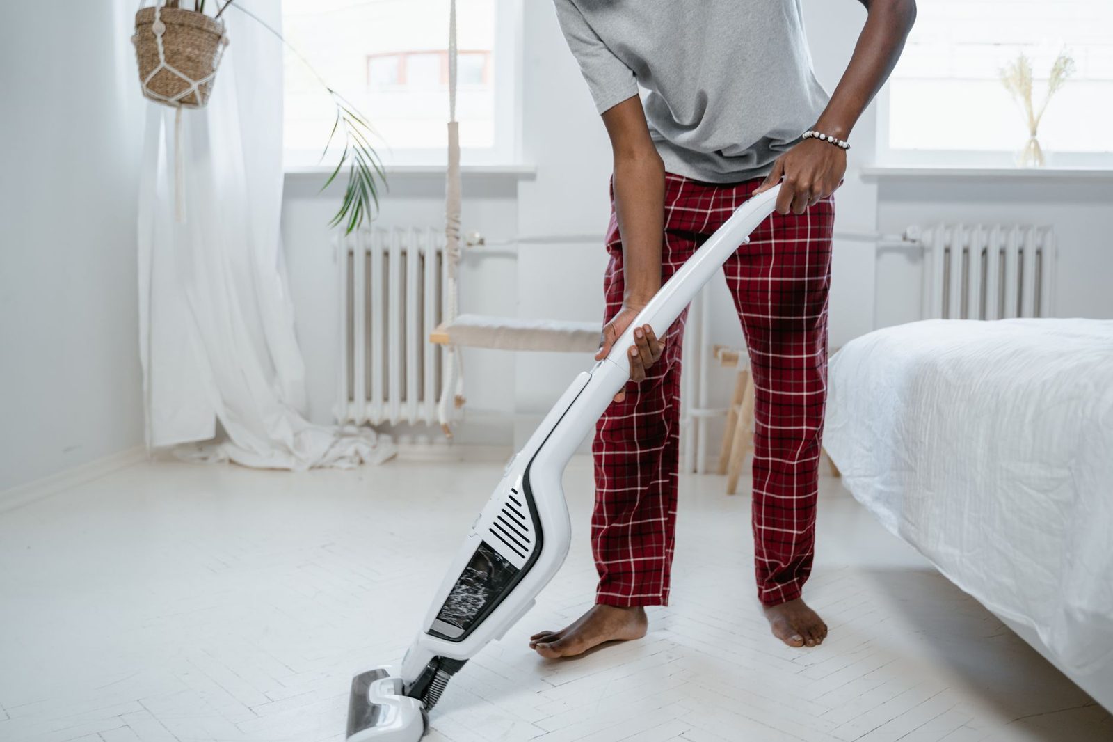 Personne en pyjama passant l'aspirateur balai sur le sol blanc d'une chambre, à côté d'un lit