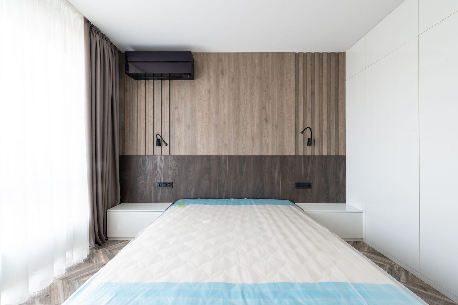 Chambre moderne avec matelas nu sur le lit, tête de lit en bois, liseuses et climatiseur mural