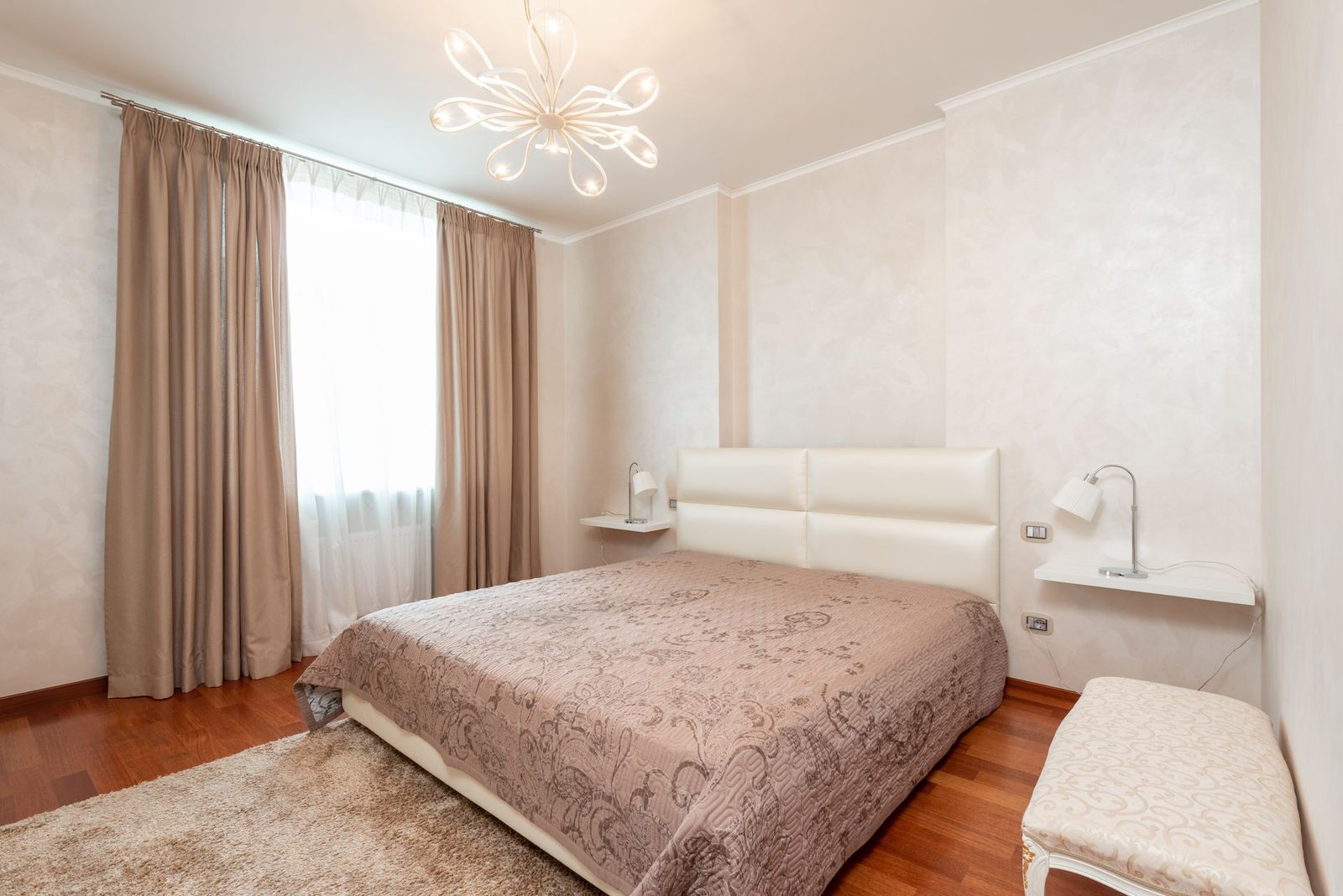 Helles Schlafzimmer mit Doppelbett, beiger Tagesdecke, Vorhängen und Holzparkett