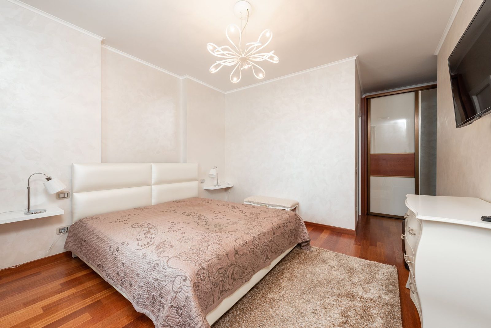 Helles Schlafzimmer mit Doppelbett, rosafarbener Tagesdecke, Parkett, Teppich und weißer Kommode