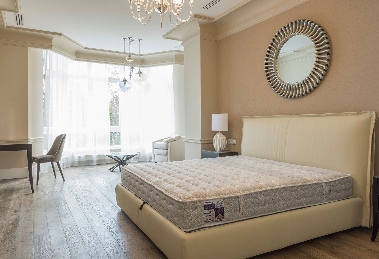 Chambre claire avec matelas nu sur un lit beige, miroir rond, lustre et parquet en bois