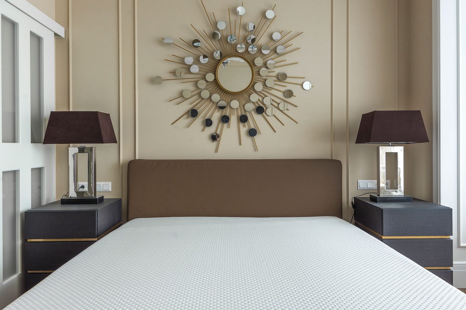 Slaapkamer met bed met witte matras, bruin hoofdeinde, nachtkastjes, lampen en zonspiegel