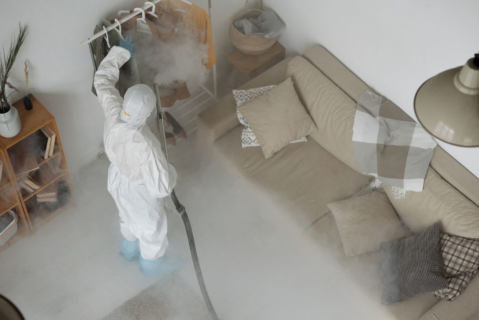 Technicien en combinaison blanche traitant un salon à la vapeur, fumée près du canapé et de l'étagère